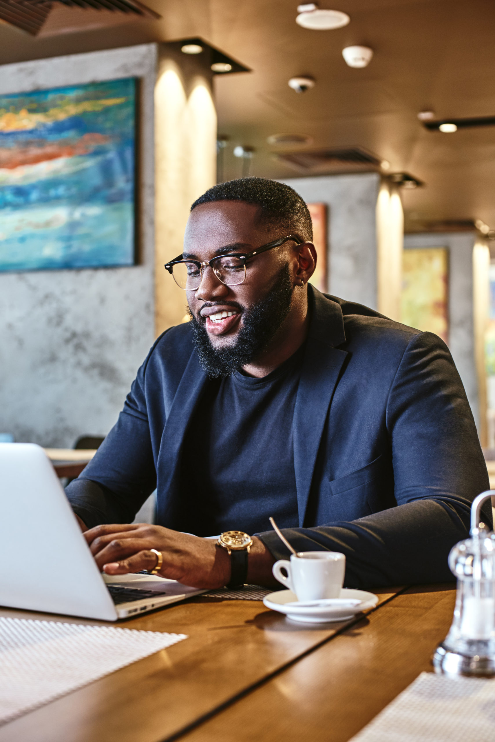 Portrait of dark-skinned businessman in dark-blue jacket and glasses sitting at the table, indoors, smiling, working with his laptop computer, drinking morning coffee. Young African entrepreneur waiting for someone in the cafe. Modern interior